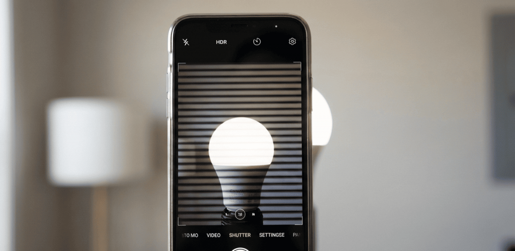 A close-up photo of a smartphone screen holding up a camera app to a bright light bulb. On the smartphone screen, distinct horizontal black banding lines are visible, illustrating the "flicker" effect of cheap LEDs. The background is slightly blurred. 